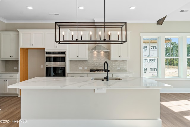 kitchen with stainless steel double oven, white cabinetry, pendant lighting, light stone countertops, and a kitchen island with sink