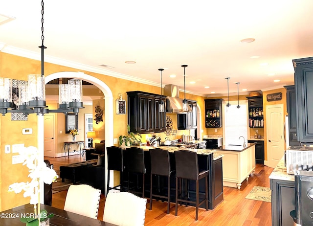 kitchen featuring light hardwood / wood-style flooring, wall chimney exhaust hood, a kitchen island with sink, a kitchen breakfast bar, and ornamental molding