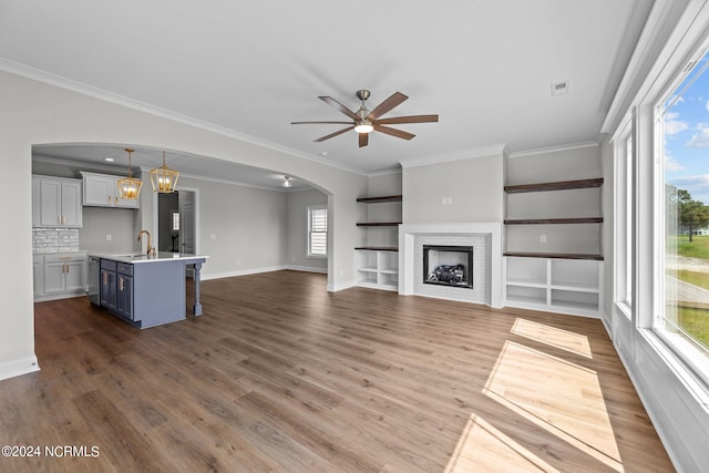 unfurnished living room featuring a brick fireplace, hardwood / wood-style floors, ceiling fan with notable chandelier, sink, and ornamental molding