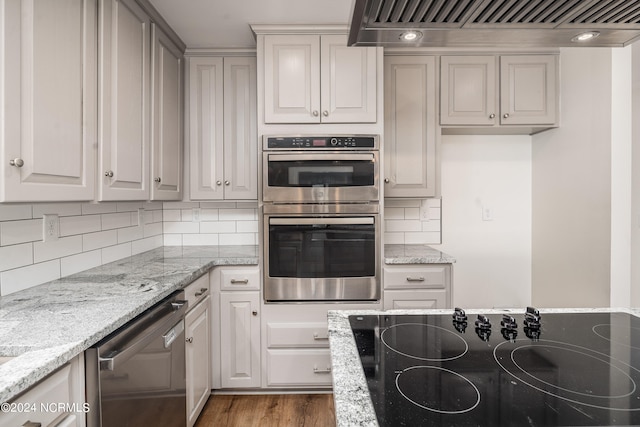 kitchen with decorative backsplash, exhaust hood, hardwood / wood-style floors, light stone countertops, and appliances with stainless steel finishes