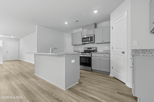 kitchen featuring gray cabinets, light stone counters, a kitchen island with sink, and appliances with stainless steel finishes