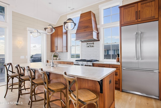 kitchen with light hardwood / wood-style flooring, an island with sink, stainless steel built in fridge, pendant lighting, and premium range hood