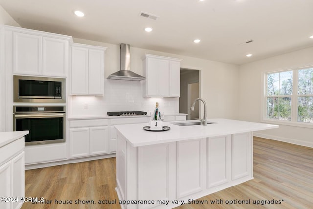 kitchen featuring built in microwave, sink, oven, a center island with sink, and wall chimney exhaust hood