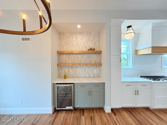 bar featuring light wood-type flooring, custom exhaust hood, stainless steel gas cooktop, beverage cooler, and white cabinets