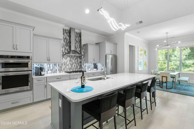 kitchen with an inviting chandelier, wall chimney range hood, sink, an island with sink, and stainless steel appliances