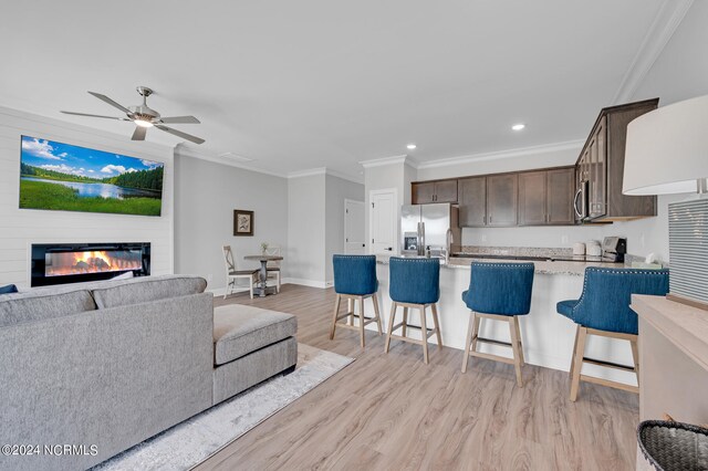 living area featuring a glass covered fireplace, baseboards, ornamental molding, light wood-style flooring, and a ceiling fan