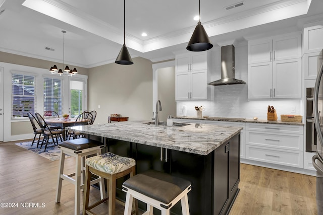 kitchen with light wood-style flooring, a sink, visible vents, wall chimney range hood, and a raised ceiling