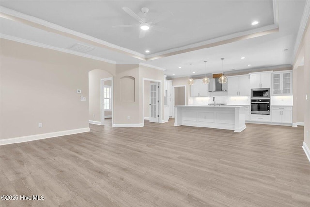 unfurnished living room with arched walkways, light wood-style flooring, a sink, ornamental molding, and a tray ceiling