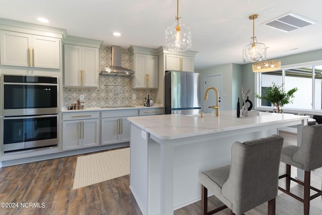 kitchen featuring wall chimney exhaust hood, dark wood-type flooring, tasteful backsplash, a center island with sink, and appliances with stainless steel finishes