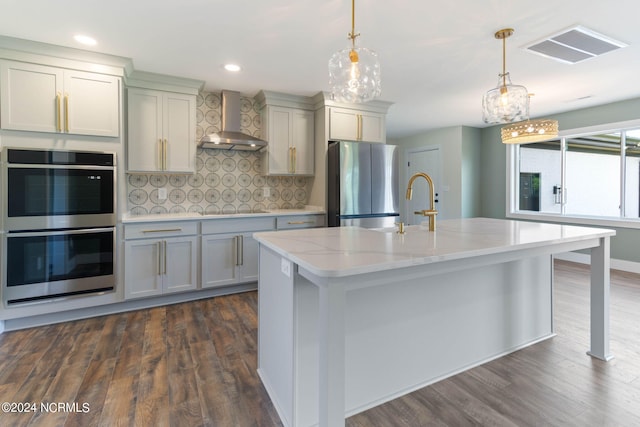 kitchen featuring wall chimney range hood, light stone counters, backsplash, an island with sink, and appliances with stainless steel finishes