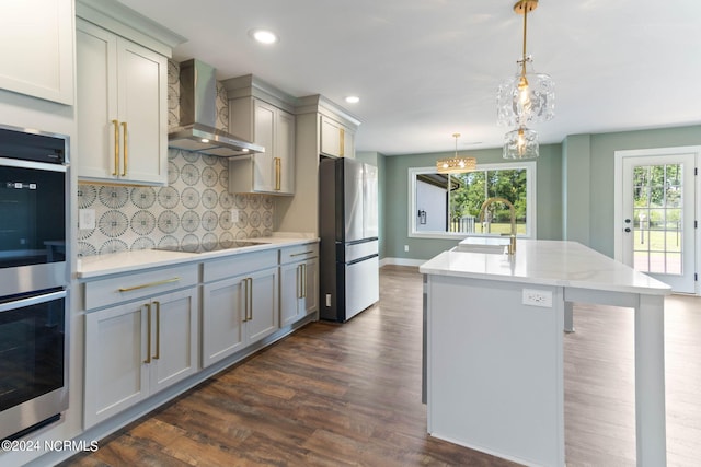 kitchen featuring wall chimney exhaust hood, stainless steel appliances, backsplash, pendant lighting, and a center island with sink