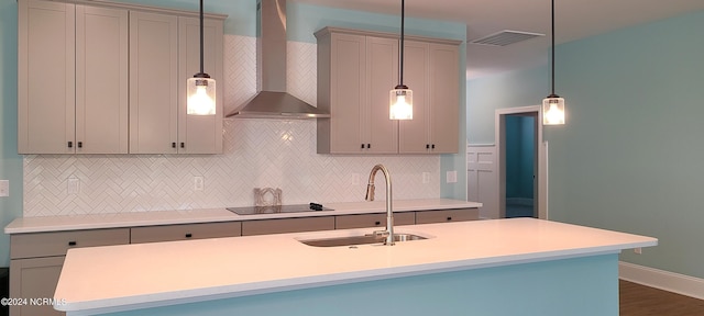 kitchen with sink, a center island with sink, black electric cooktop, pendant lighting, and wall chimney range hood