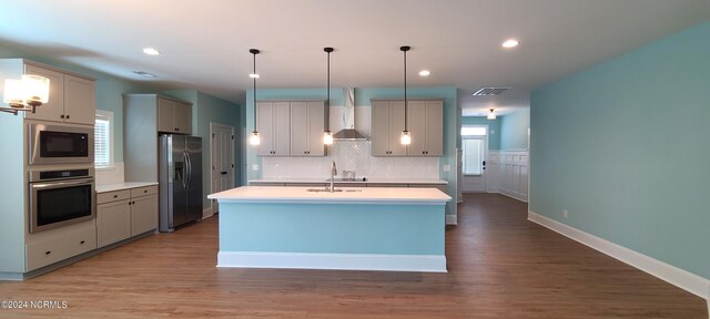 kitchen with wall chimney range hood, hardwood / wood-style flooring, stainless steel appliances, a center island with sink, and pendant lighting