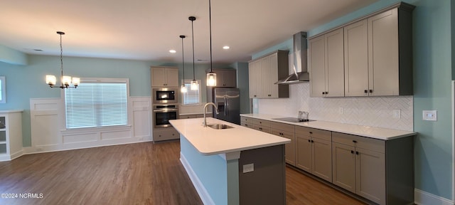 kitchen with stainless steel appliances, decorative light fixtures, an island with sink, and wall chimney range hood