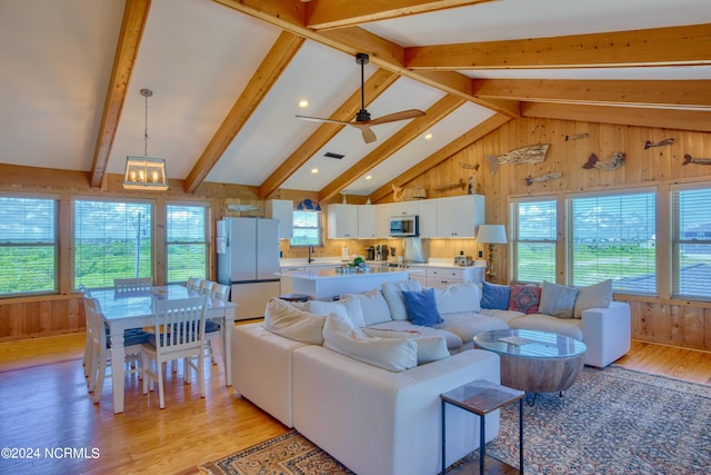 living room with wood walls, ceiling fan with notable chandelier, sink, vaulted ceiling with beams, and light wood-type flooring