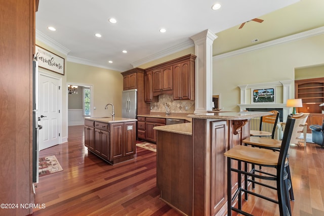 kitchen with sink, ornamental molding, an island with sink, appliances with stainless steel finishes, and decorative columns