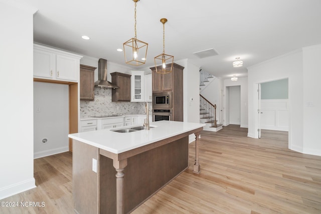 kitchen with wall chimney range hood, white cabinets, stainless steel appliances, and a center island with sink