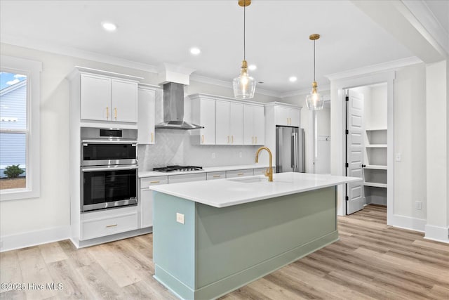 kitchen featuring an island with sink, sink, white cabinets, stainless steel appliances, and wall chimney range hood