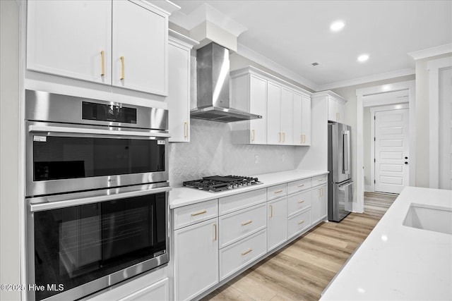 kitchen with appliances with stainless steel finishes, white cabinetry, ornamental molding, wall chimney exhaust hood, and light wood-type flooring