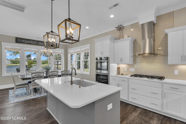 kitchen with wall chimney range hood, appliances with stainless steel finishes, an island with sink, white cabinets, and decorative light fixtures