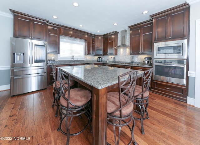 kitchen featuring appliances with stainless steel finishes, wall chimney range hood, dark brown cabinets, a center island, and dark hardwood / wood-style flooring