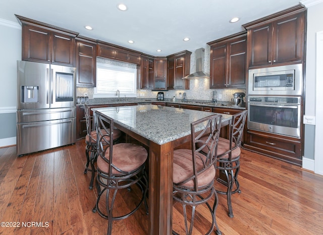 kitchen with tasteful backsplash, wall chimney exhaust hood, a kitchen island, appliances with stainless steel finishes, and light stone counters