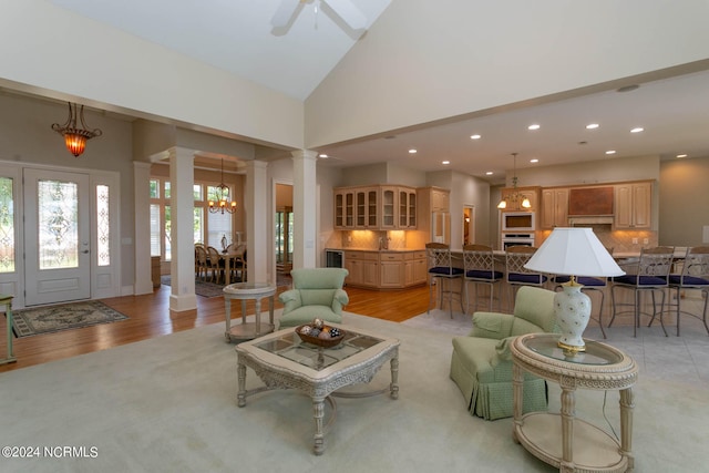 living room with ceiling fan with notable chandelier, ornate columns, light wood-type flooring, and high vaulted ceiling