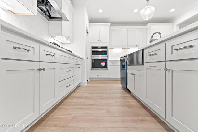 kitchen featuring stainless steel double oven, white cabinets, light countertops, wall chimney exhaust hood, and pendant lighting