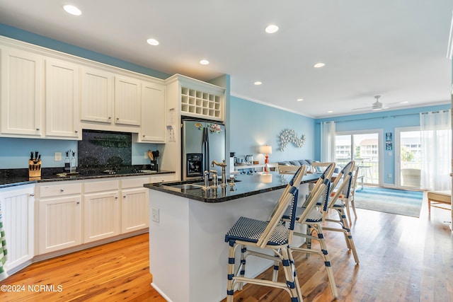 kitchen featuring stainless steel fridge with ice dispenser, light hardwood / wood-style flooring, dark stone counters, a kitchen island with sink, and ceiling fan