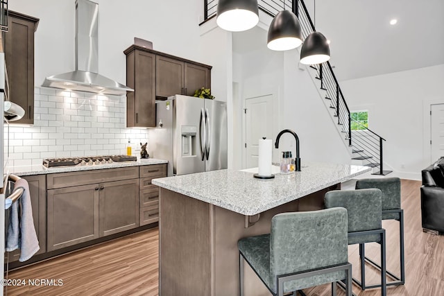 kitchen featuring wall chimney range hood, light hardwood / wood-style flooring, stainless steel appliances, light stone counters, and an island with sink