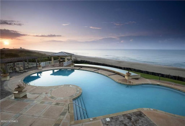 pool at dusk featuring a gazebo, a water view, a beach view, and a patio area