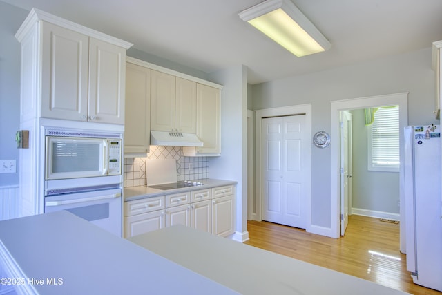 kitchen with white appliances, light wood-style floors, light countertops, under cabinet range hood, and backsplash