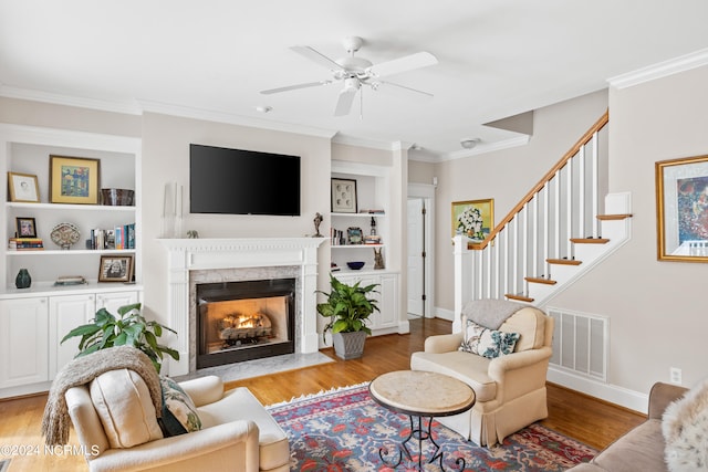 living room with a high end fireplace, light hardwood / wood-style floors, built in shelves, ornamental molding, and ceiling fan