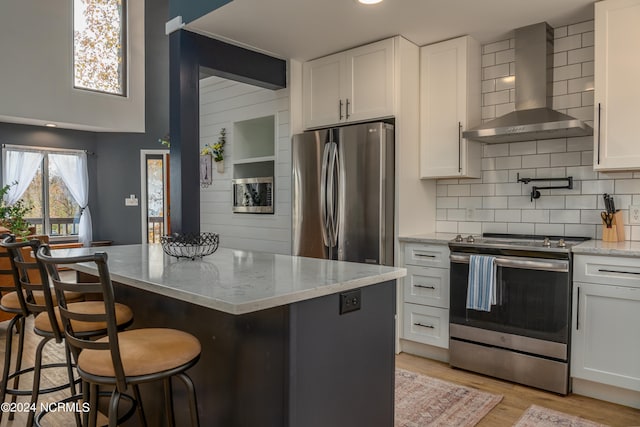 kitchen featuring wall chimney exhaust hood, stainless steel appliances, light stone counters, light wood-type flooring, and white cabinets