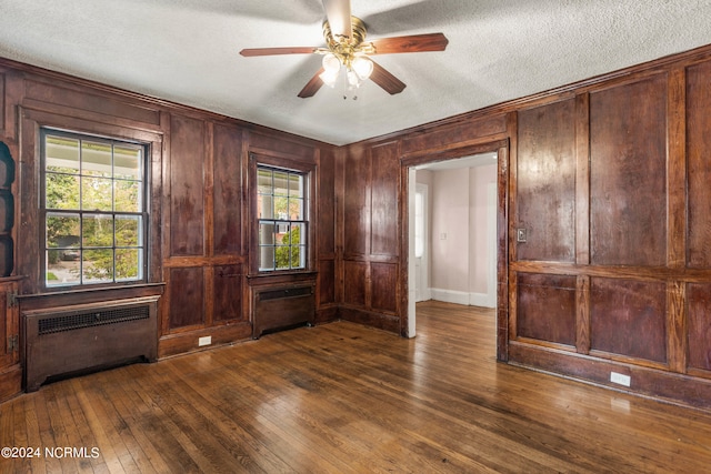 unfurnished room with wooden walls, radiator heating unit, dark wood-type flooring, a textured ceiling, and ceiling fan