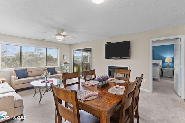 dining room featuring light carpet, baseboards, a ceiling fan, a glass covered fireplace, and a textured ceiling