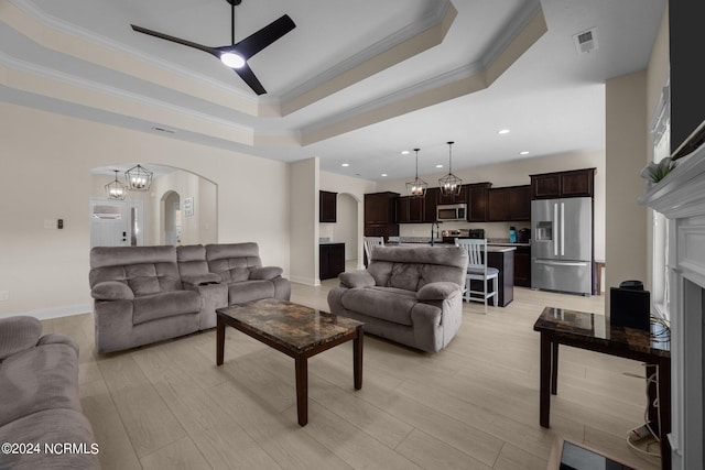 living room with a tray ceiling, ornamental molding, ceiling fan with notable chandelier, light hardwood / wood-style floors, and sink