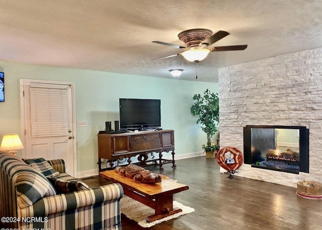 living area featuring a textured ceiling, wood finished floors, ceiling fan, baseboards, and a stone fireplace