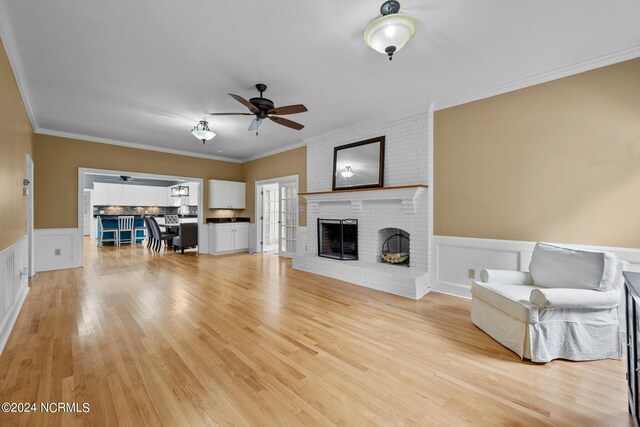 unfurnished living room with a wainscoted wall, ornamental molding, a ceiling fan, a brick fireplace, and light wood-type flooring