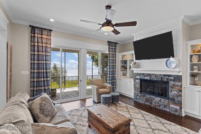 living room with built in features, a stone fireplace, dark wood-type flooring, ceiling fan, and ornamental molding