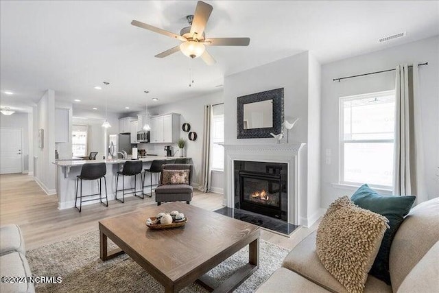 living area featuring light wood finished floors, visible vents, a glass covered fireplace, ceiling fan, and baseboards