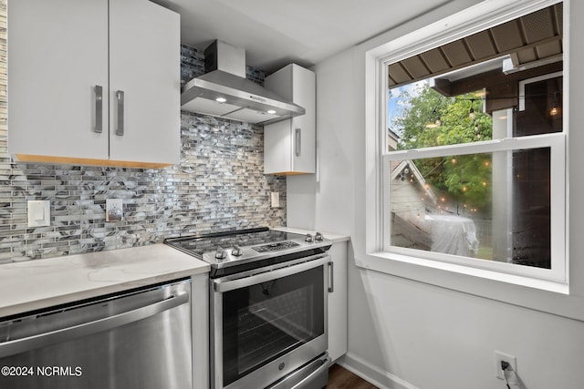 kitchen featuring appliances with stainless steel finishes, tasteful backsplash, white cabinetry, hardwood / wood-style flooring, and wall chimney range hood