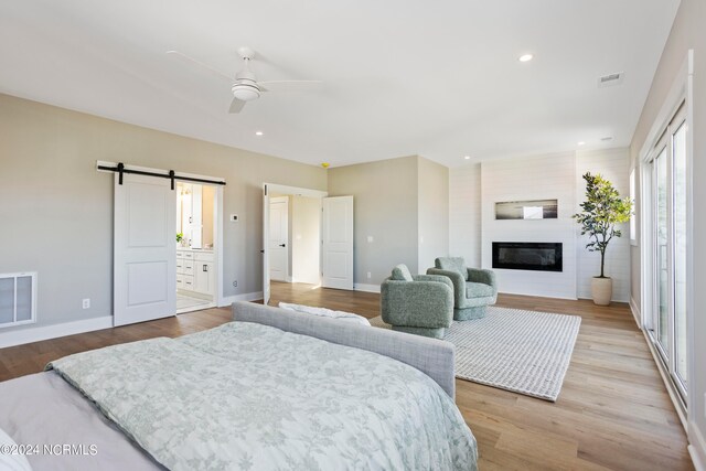 bedroom featuring light wood-type flooring, a barn door, ensuite bath, a large fireplace, and ceiling fan