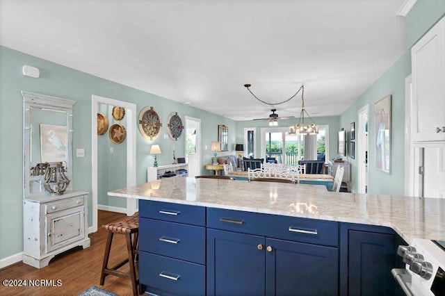 kitchen featuring dark wood-type flooring, ceiling fan with notable chandelier, blue cabinetry, light stone counters, and a kitchen bar