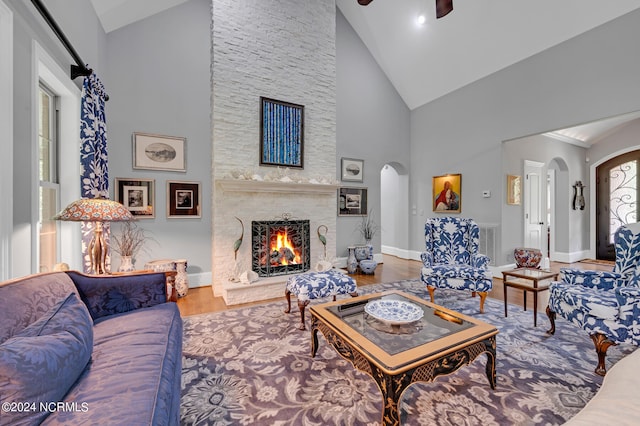 living room with arched walkways, a stone fireplace, wood finished floors, a ceiling fan, and baseboards