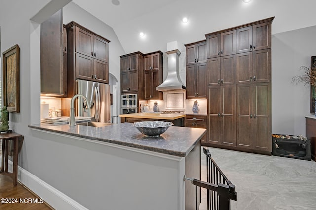 kitchen featuring decorative backsplash, vaulted ceiling, stainless steel appliances, dark brown cabinets, and wall chimney range hood