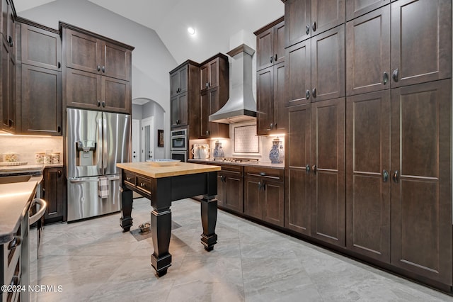 kitchen featuring arched walkways, dark brown cabinetry, vaulted ceiling, appliances with stainless steel finishes, and wall chimney range hood