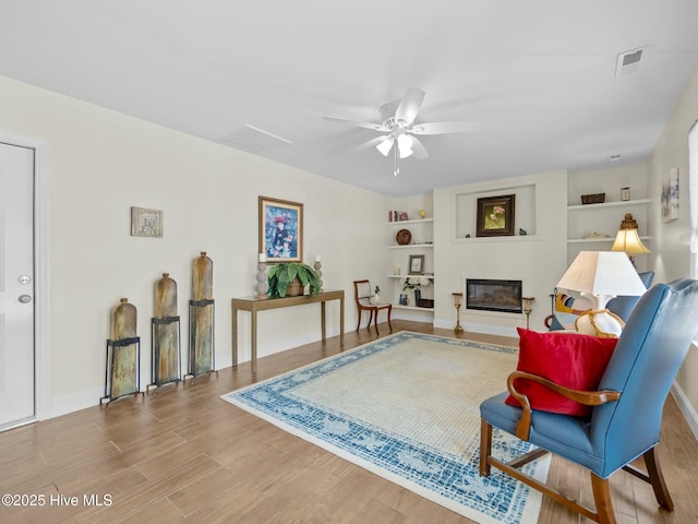 sitting room featuring built in features, wood finished floors, visible vents, ceiling fan, and a glass covered fireplace