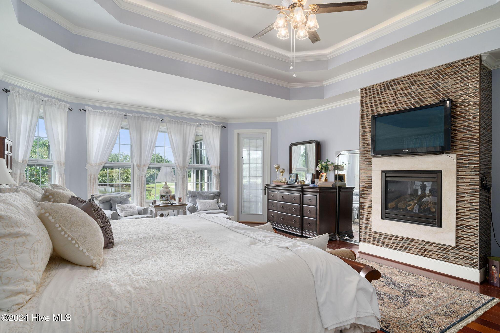 bedroom featuring ceiling fan, a large fireplace, crown molding, wood-type flooring, and a tray ceiling