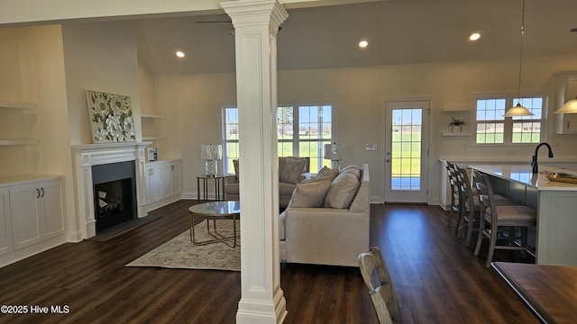 living area featuring dark wood-type flooring, a fireplace with raised hearth, and ornate columns